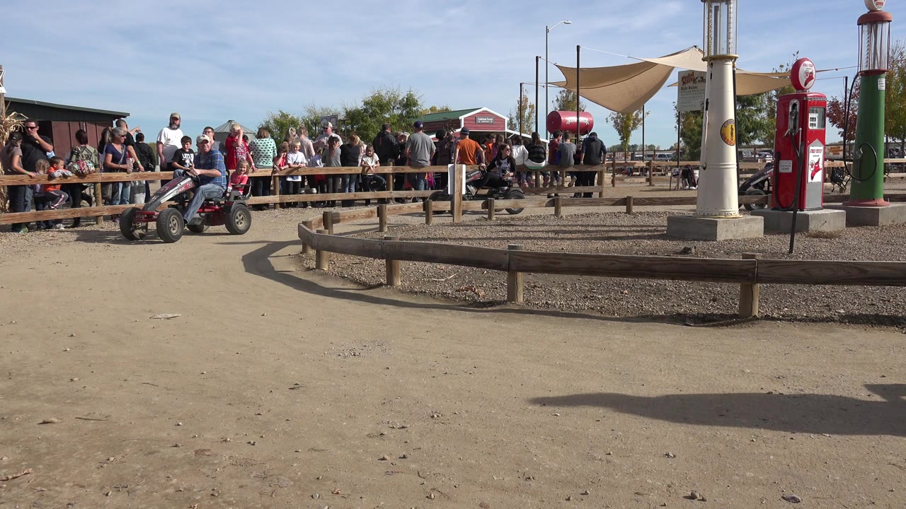 A pedal go-kart rider moves near the left edge of an outdoor dirt track in Brighton, Colorado, with spectators behind a wooden fence and pump-like structures in the background.
