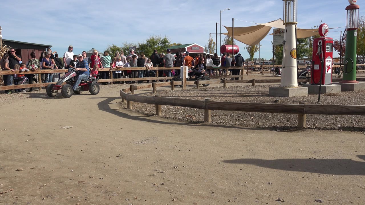 A wide view of the Brighton, Colorado kart area shows riders entering from the left, a low wood rail in the foreground, and a group of spectators gathered behind the fence.