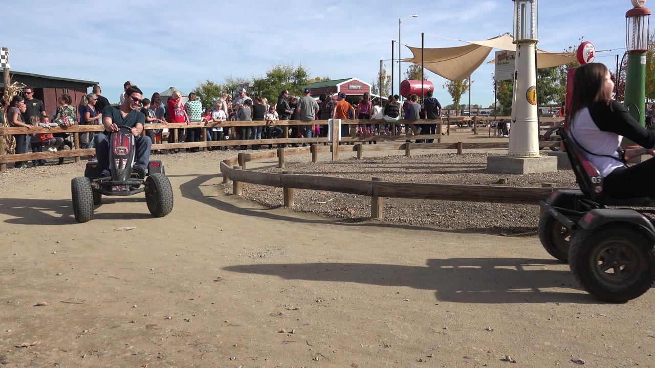 At a Brighton, Colorado public event, a child on a low pedal vehicle rides near the left side of a dirt circle while people watch from behind a wooden rail.
