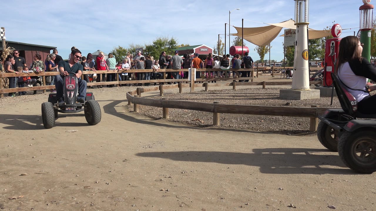 A foreground rider on a small pedal vehicle crosses the frame while another child rides toward the camera on the dirt circle at the Brighton, Colorado event.