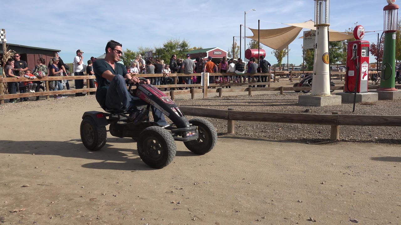 Adult rider crosses the dirt circle on a pedal vehicle in front of a crowd and red pump decor.