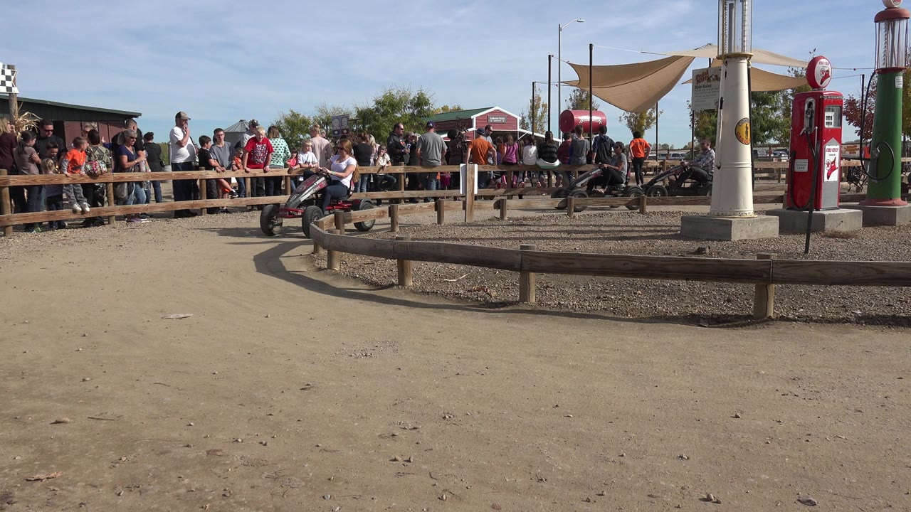 Adults on pedal tractor carts approach a dirt track corner in front of spectators.