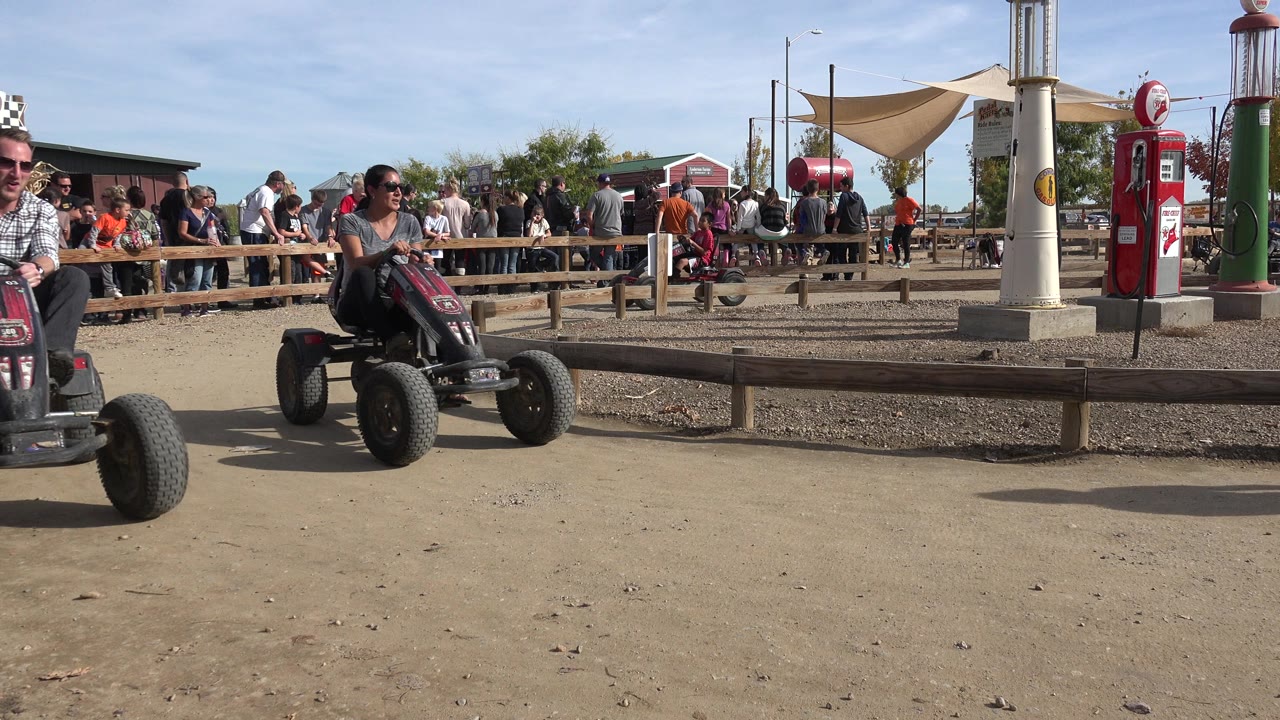 At the start of the clip in Brighton, Colorado, riders on four-wheel pedal carts move along a dirt track while a crowd watches behind a wooden rail.