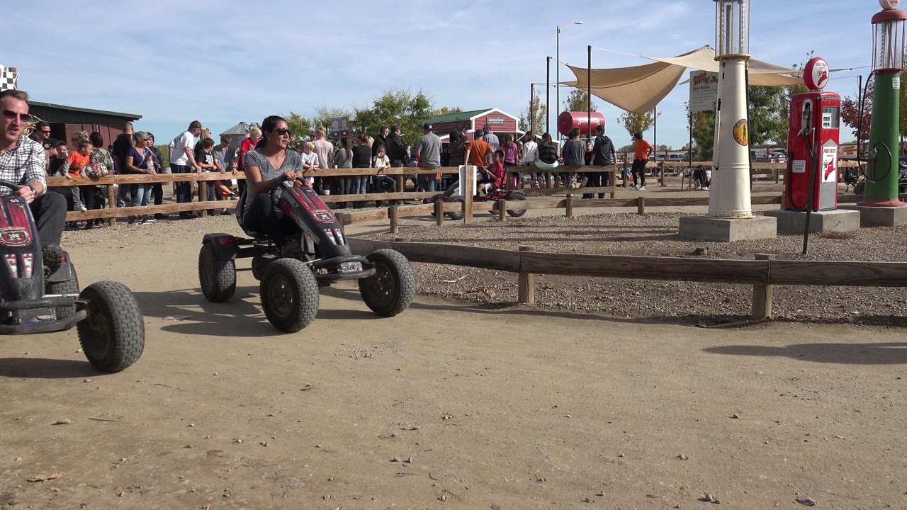 A four-wheel pedal cart approaches the camera in Brighton, Colorado, with spectators, shade sails, and red fuel-pump-style props visible behind the track.