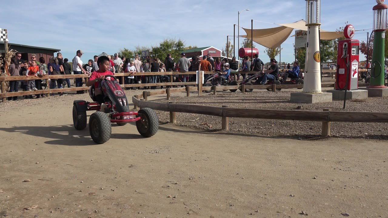 Boy in a red shirt drives a pedal go-kart on a dirt track beside spectators.