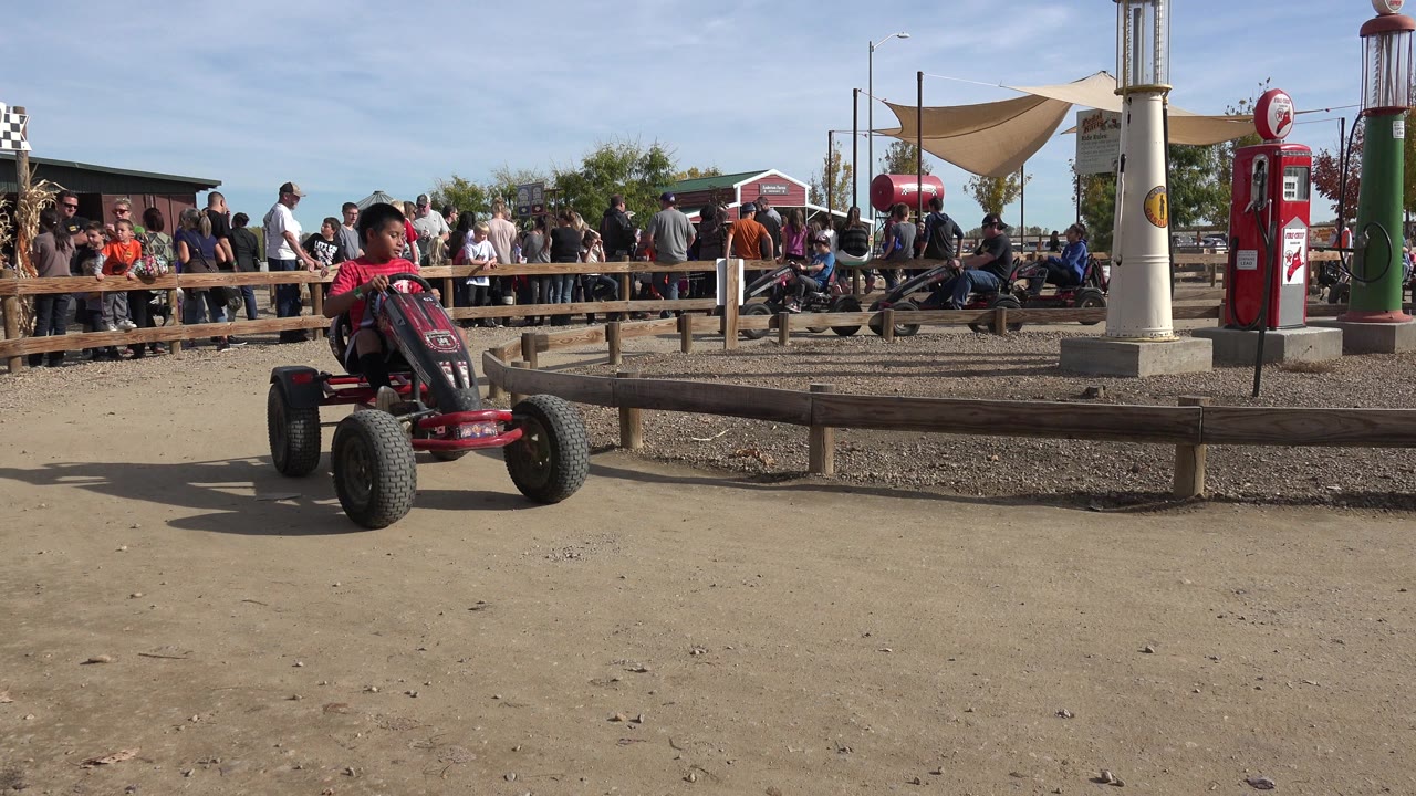 At 0:01 in Brighton, Colorado, the red-shirted boy is farther through the corner, with the go-kart angled across the dusty foreground and the crowd watching behind him.