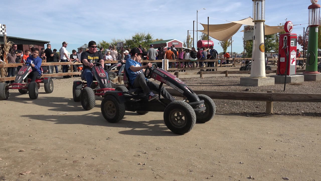 Pedal carts pass on a dusty amusement track with spectators behind a rail.