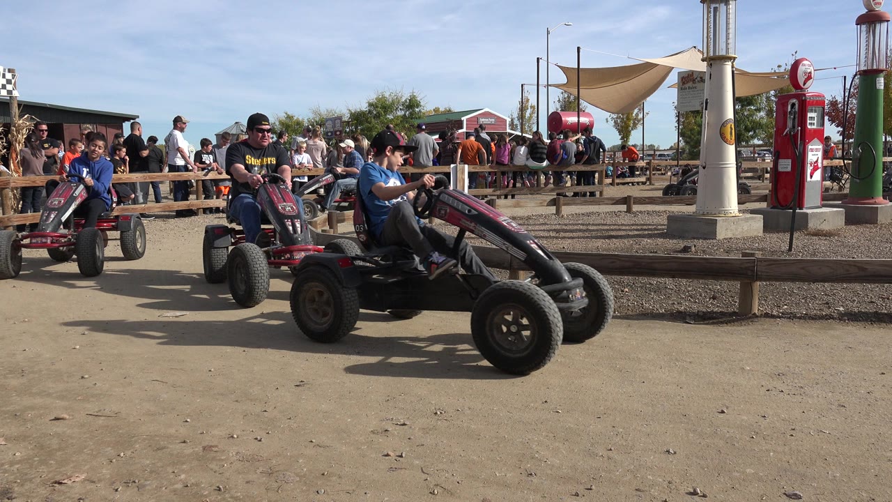 In Brighton, Colorado, a child in a blue shirt rides a red pedal cart near the foreground as another rider follows behind on the amusement track.