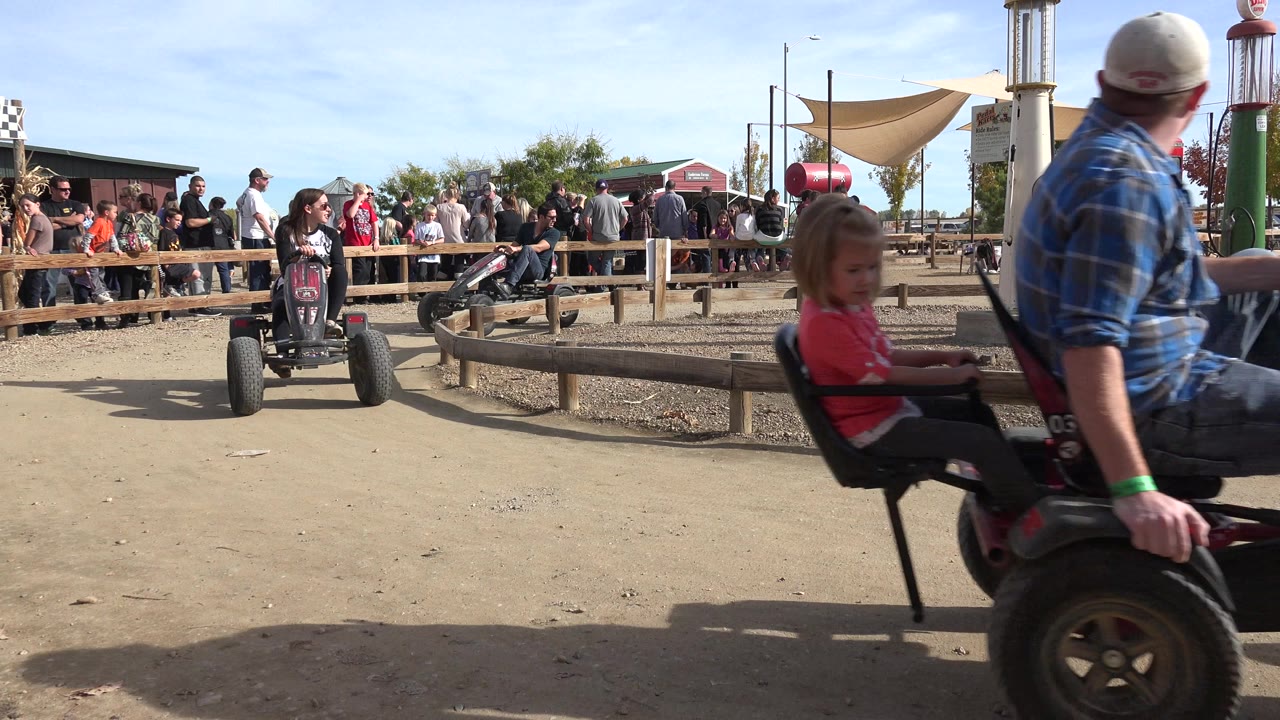 At 0:00 in Brighton, Colorado, a rider in a pedal kart approaches from the left side of a dirt track while a crowd stands behind a wooden rail under bright daylight.