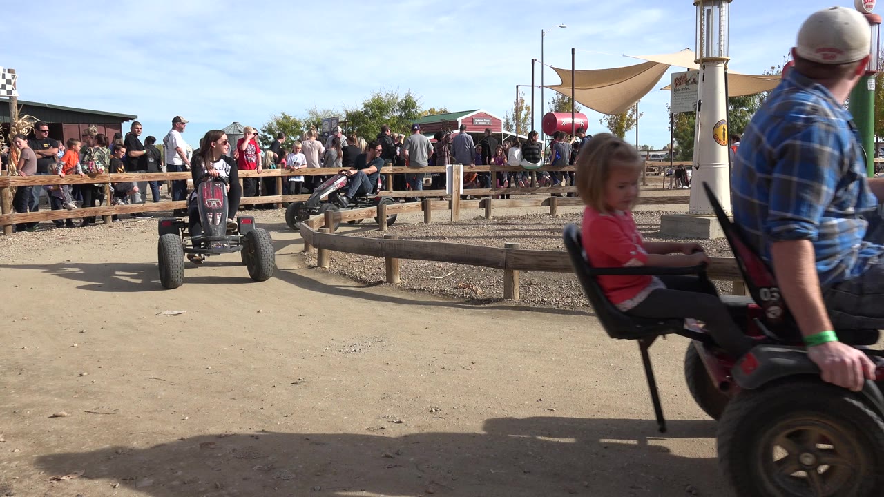 At 0:01 in Brighton, Colorado, an adult and a child in the foreground pedal kart pass close to the camera while another rider and spectators remain behind the wooden barrier.