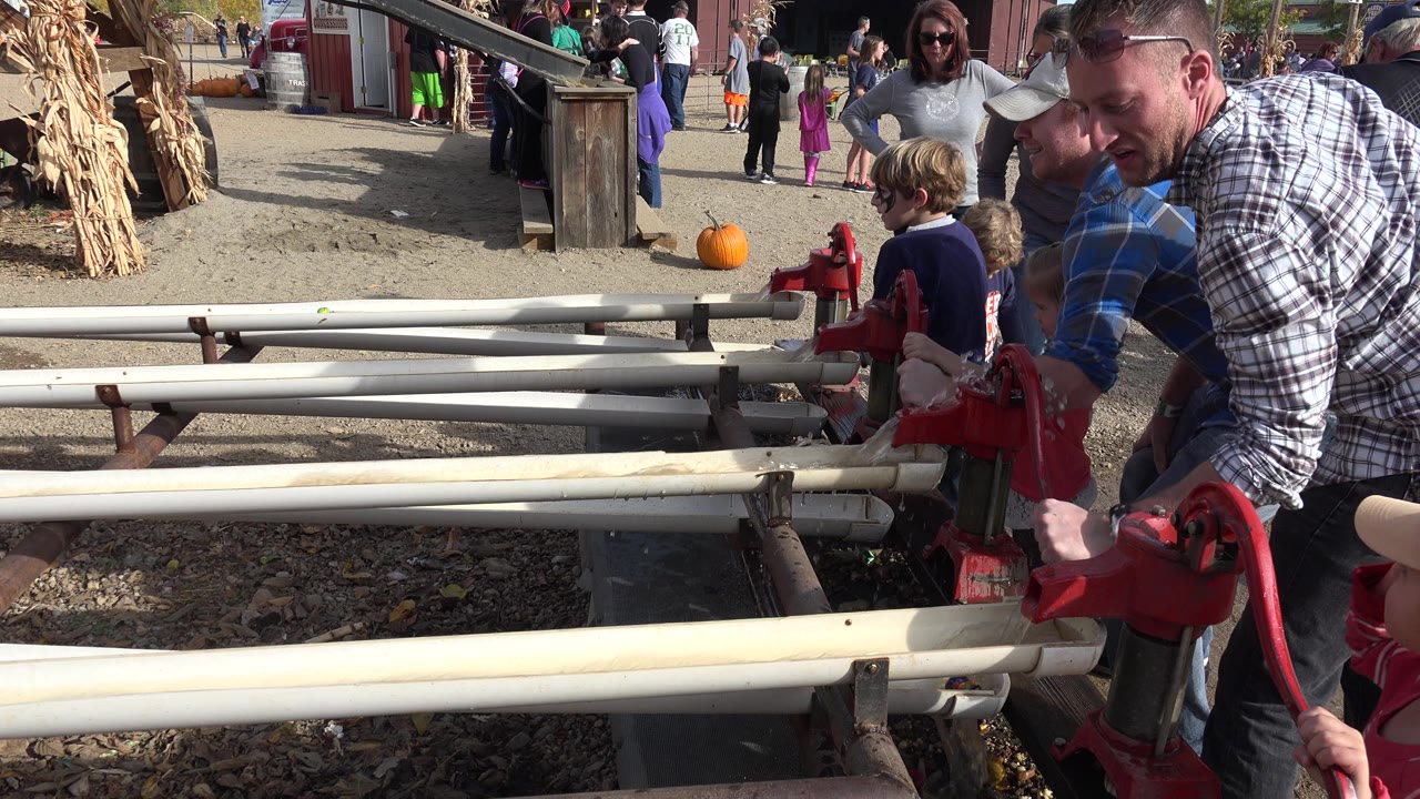 A man in a plaid shirt leans over a red hand-crank water pump in Brighton, Colorado, while children stand beside the pump rails and people move near farm buildings behind them.
