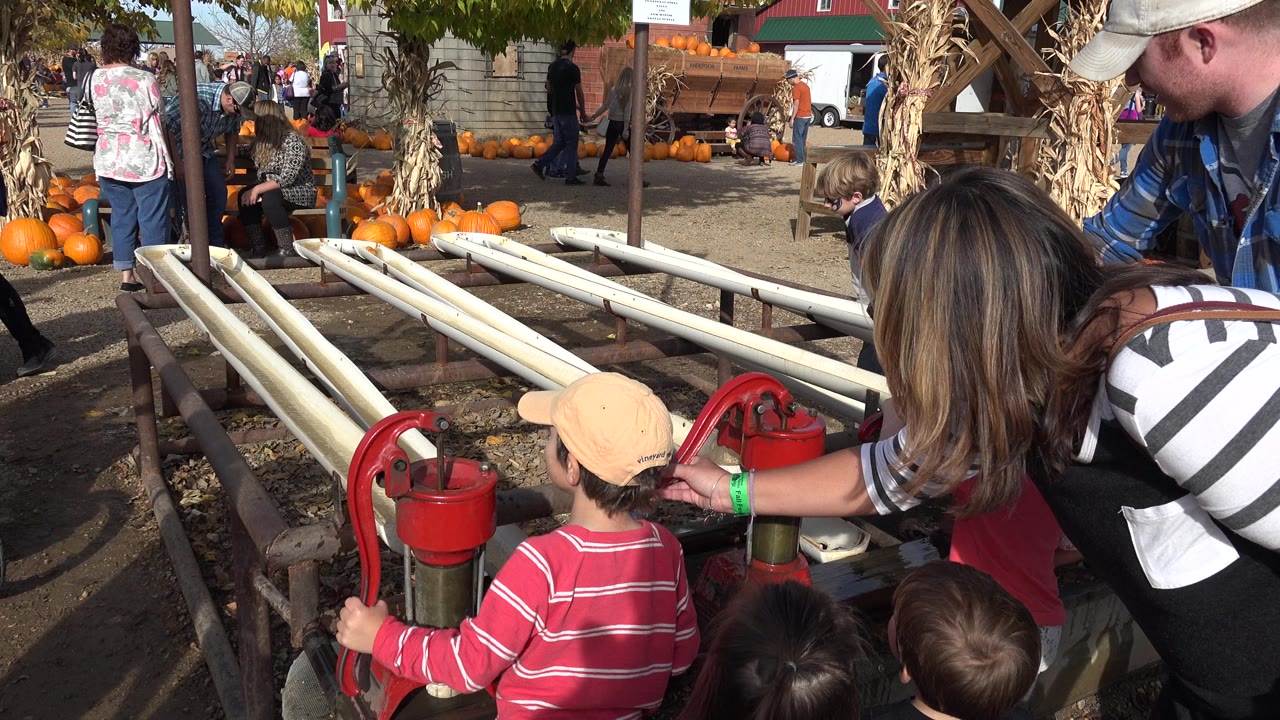 Children and adults around a red hand pump near pumpkins and rail lanes.