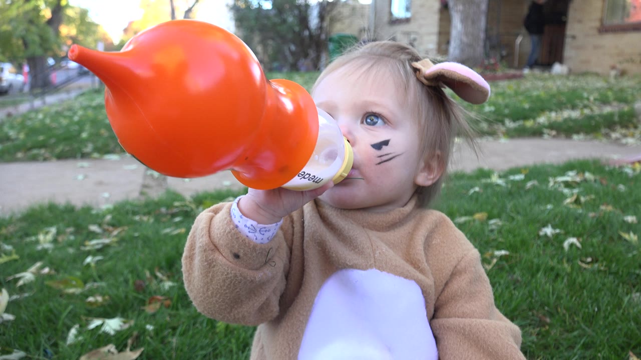 Toddler in costume drinks from bottle with orange prop raised high.