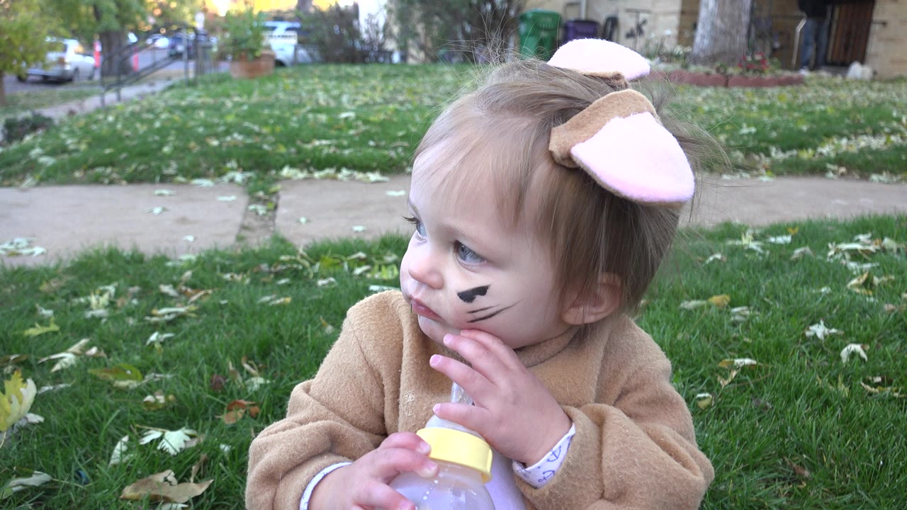 Baby in animal costume sitting on grass with a bottle.