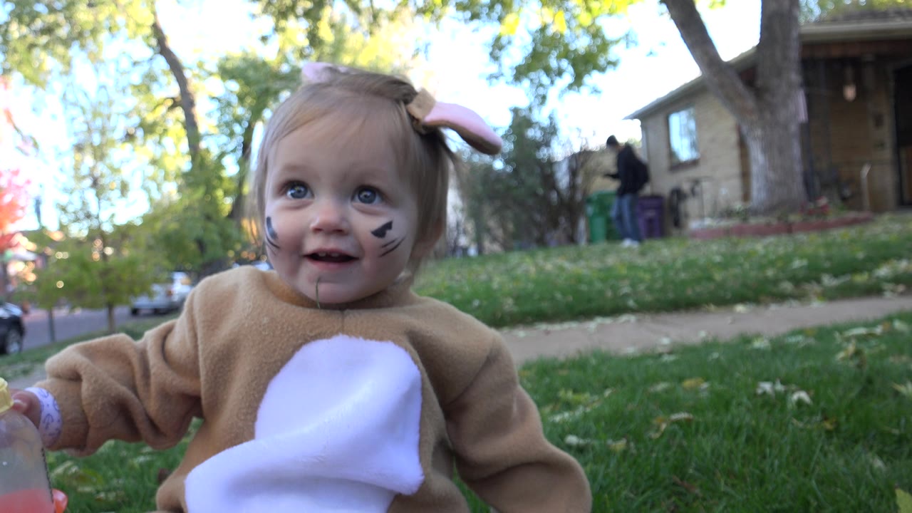 The toddler in a brown bunny costume smiles near the low camera in Westminster, Colorado, with green grass and a residential house visible in soft daylight.