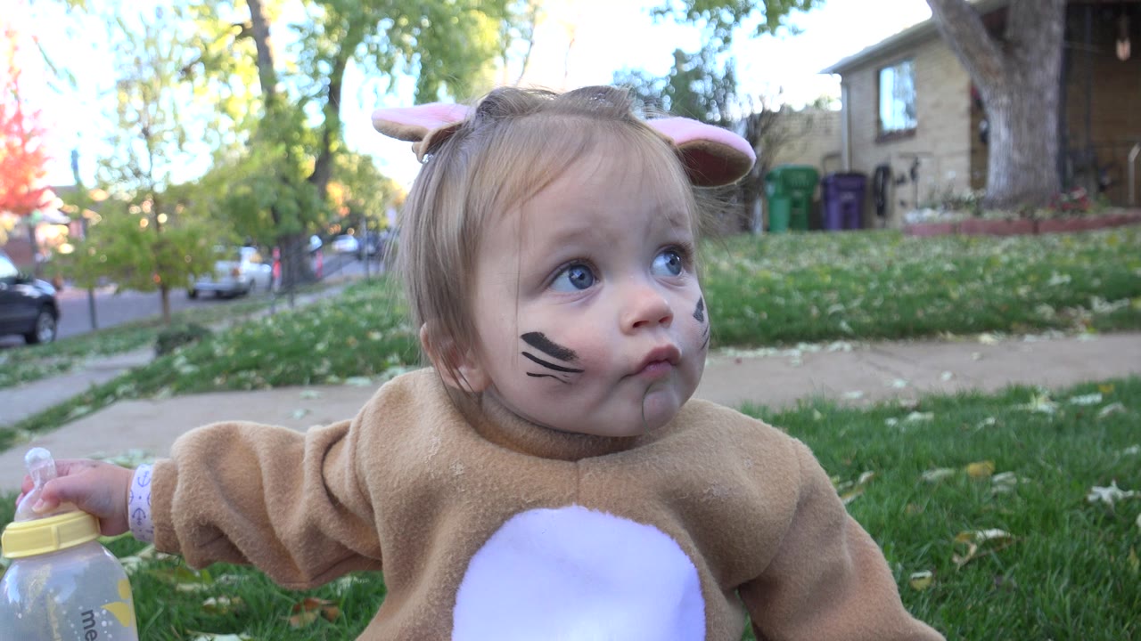 The baby remains seated on the lawn in Westminster, Colorado, looking up and to the side with face paint visible and one hand resting on the bottle cap.