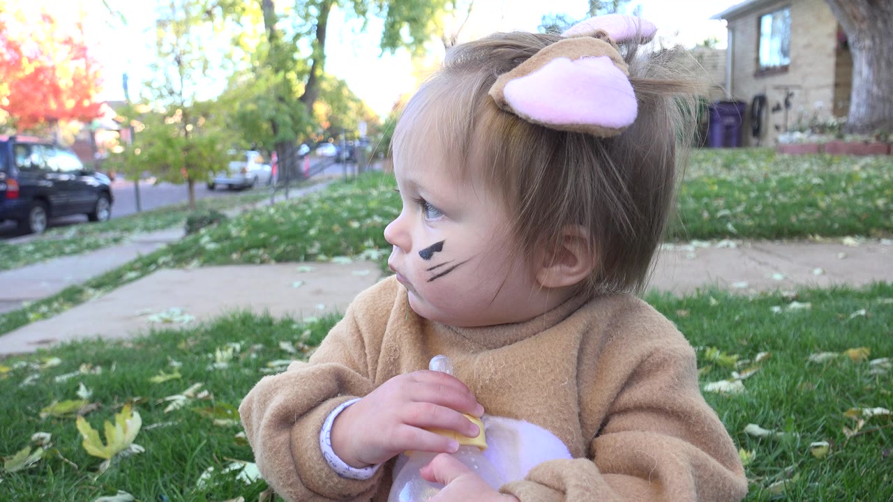 The child remains seated in the grass in Westminster, Colorado, turned left with a bottle near her chest and parked cars visible beyond the sidewalk.