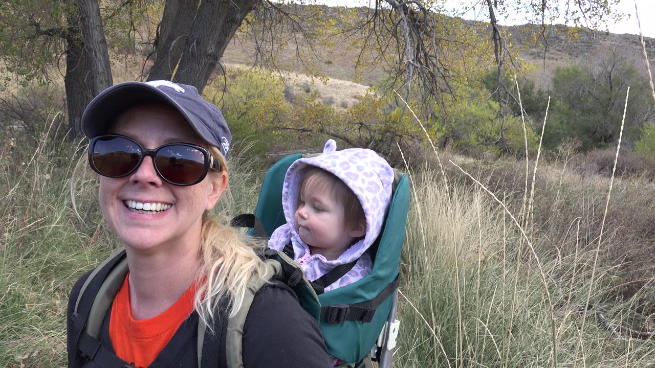 Smiling woman carries a young child in a backpack carrier on a grassy trail.