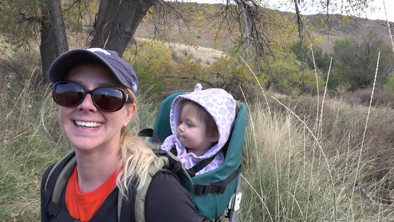 In Golden, Colorado, the woman faces the camera with a broad smile while the child turns sideways from the backpack carrier near dry grass.