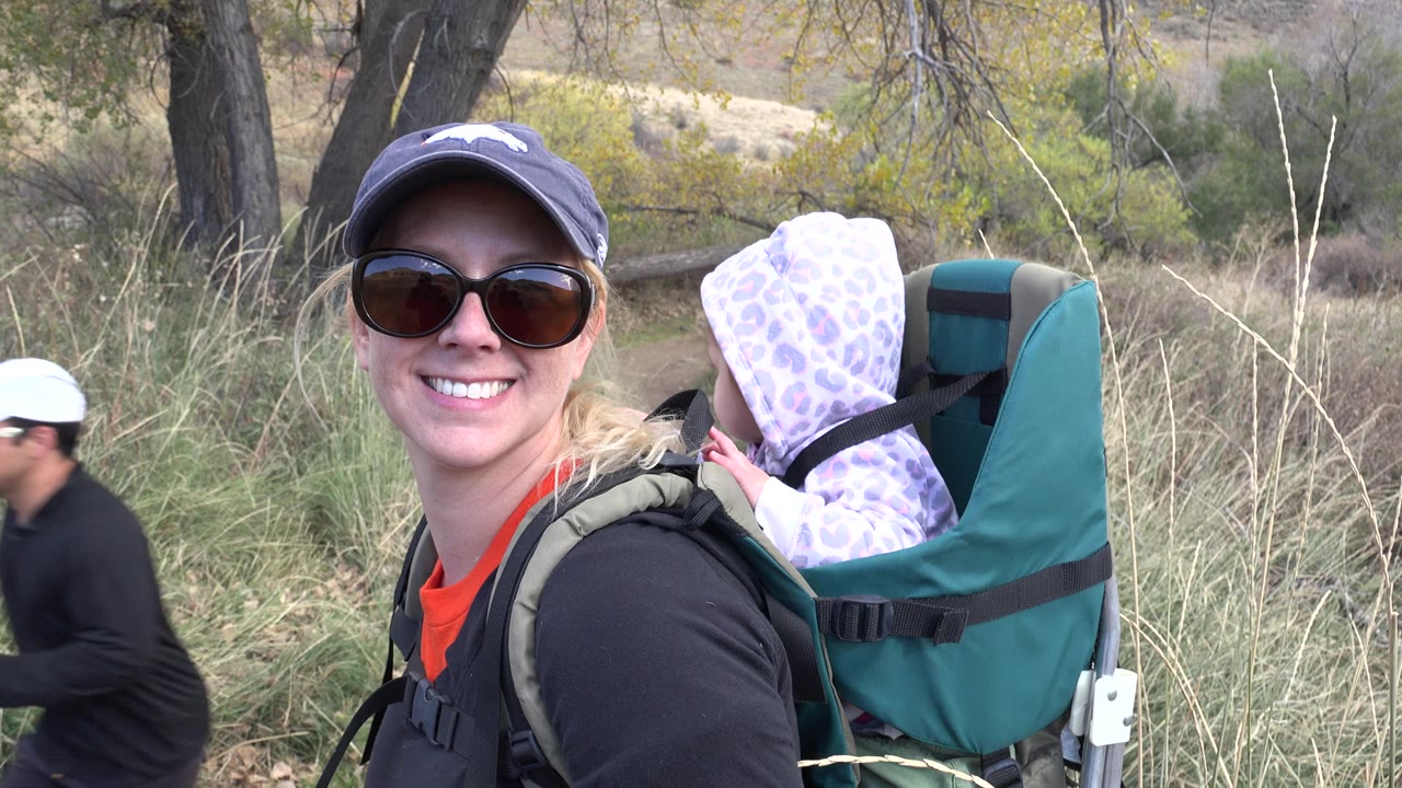 Smiling woman wearing sunglasses carries a baby backpack on a grassy trail.