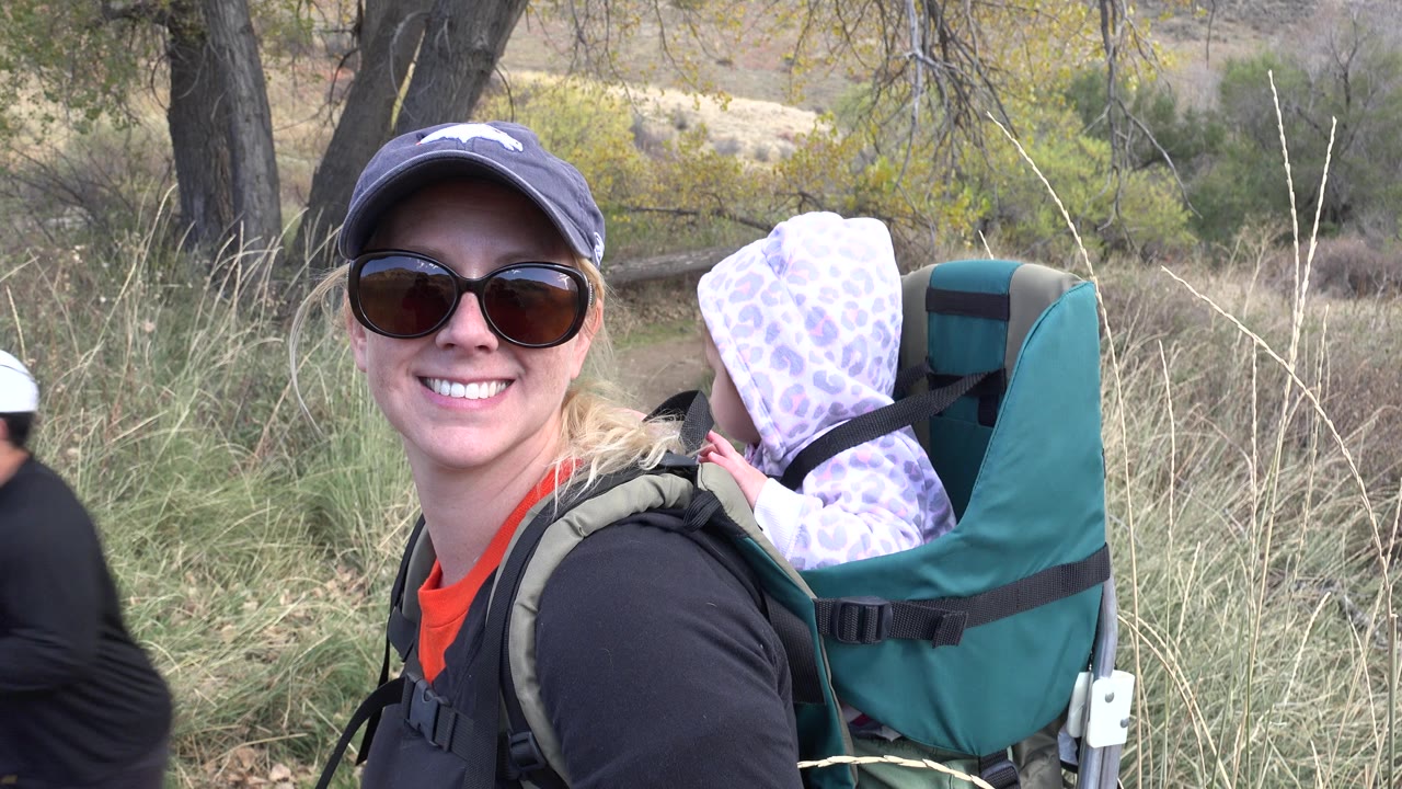 A close trail view in Golden, Colorado shows the woman smiling toward the camera with the hooded baby facing away in the green backpack carrier.