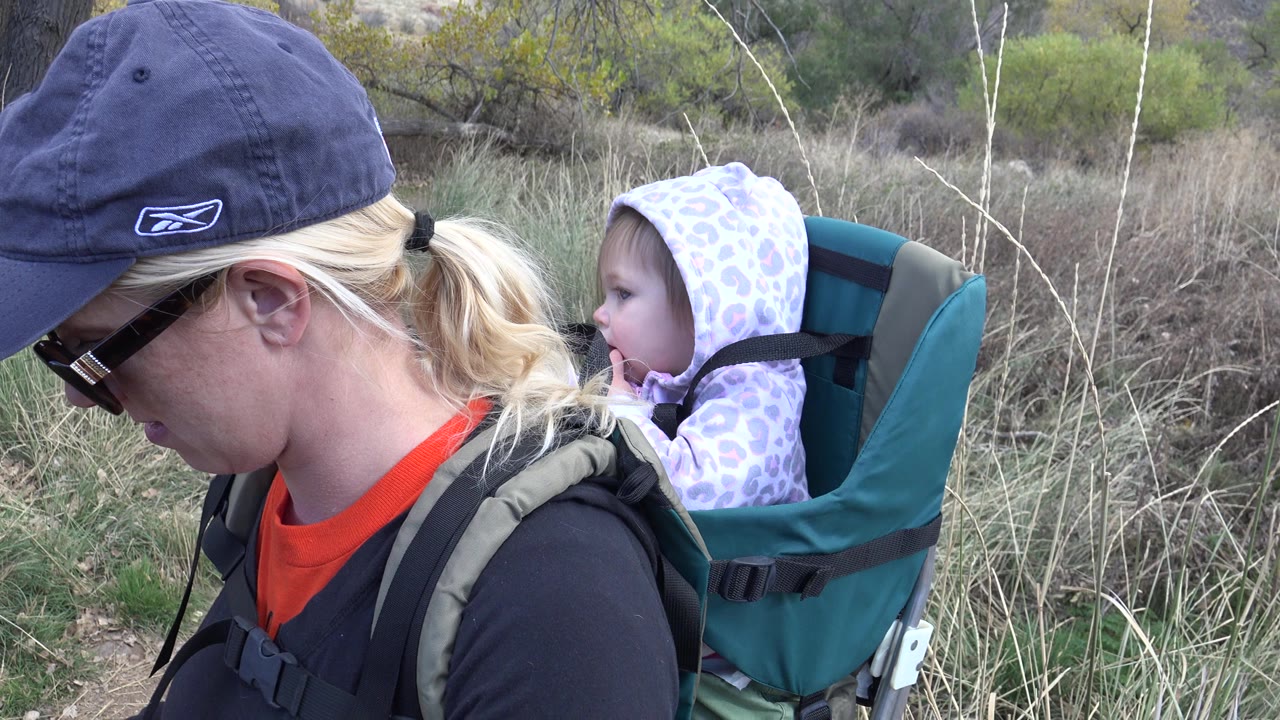 Woman wearing a cap carries a child in a backpack carrier on a trail.