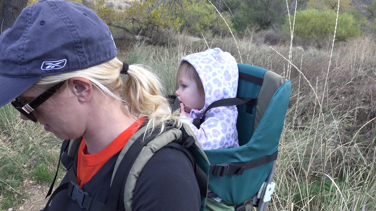 The woman remains in left profile while the child looks forward from the backpack carrier, with tall pale grasses and scrub vegetation behind them in Golden, Colorado.