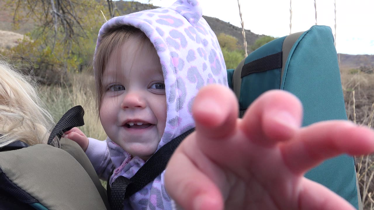 In Golden, Colorado, a toddler in a lavender hood sits in a teal backpack carrier and reaches a hand close to the camera, with dry grasses and low hills behind.