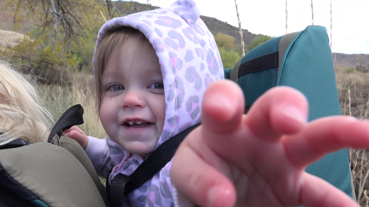 The toddler smiles from the backpack carrier with fingers extended toward the lens, while a blonde adult's hair, tall grass, and foothill terrain remain visible around the frame.