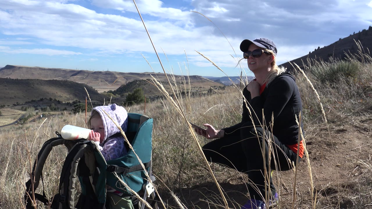 Woman beside child in a baby backpack on a sunny hillside trail.