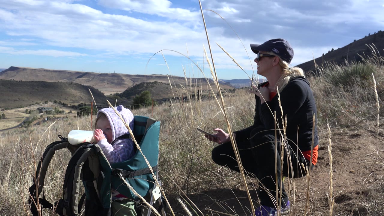Woman kneeling beside a child in a backpack carrier on a grassy hillside.