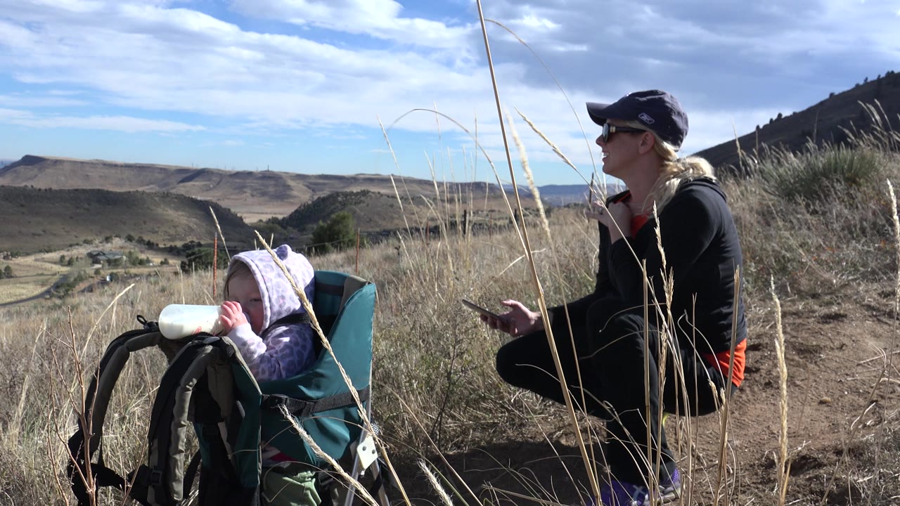A child seated in a teal hiking carrier drinks from a bottle as a woman kneels to the right, with tall grass crossing the foreground in Golden, Colorado.