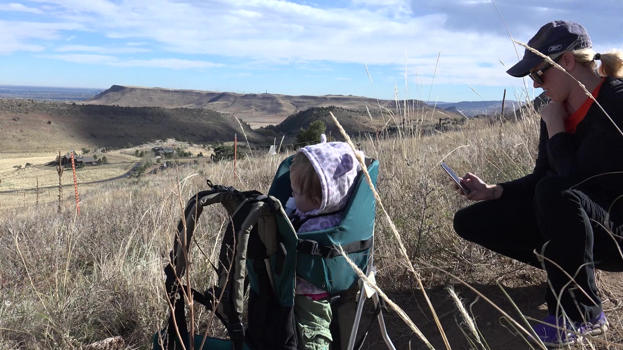 A woman kneels beside a baby in a teal backpack carrier on a grassy hillside in Golden, Colorado, with distant ridges and blue sky behind them.