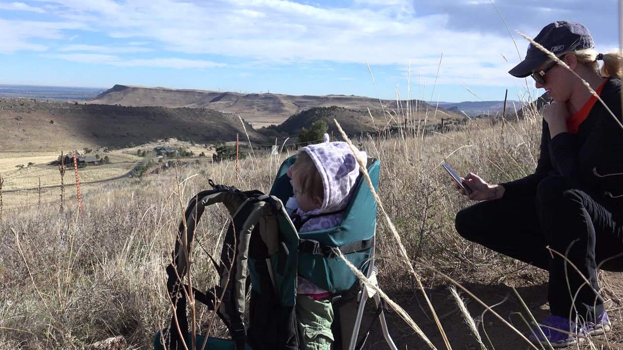 The baby sits turned toward the open hills while the mother crouches nearby looking at a phone, with tall grass crossing the foreground in Golden, Colorado.