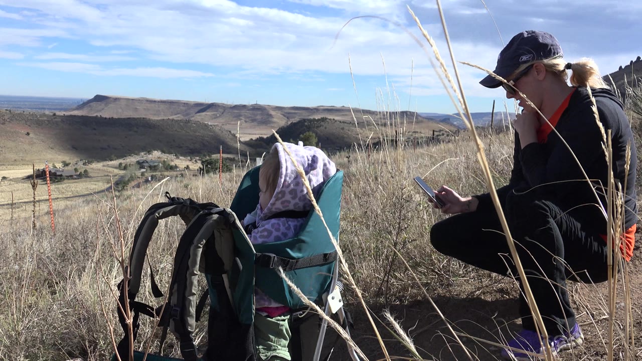 The mother stays low beside the baby carrier as both sit among dry grass, with a broad ridge line and pale sky visible across the Golden, Colorado background.