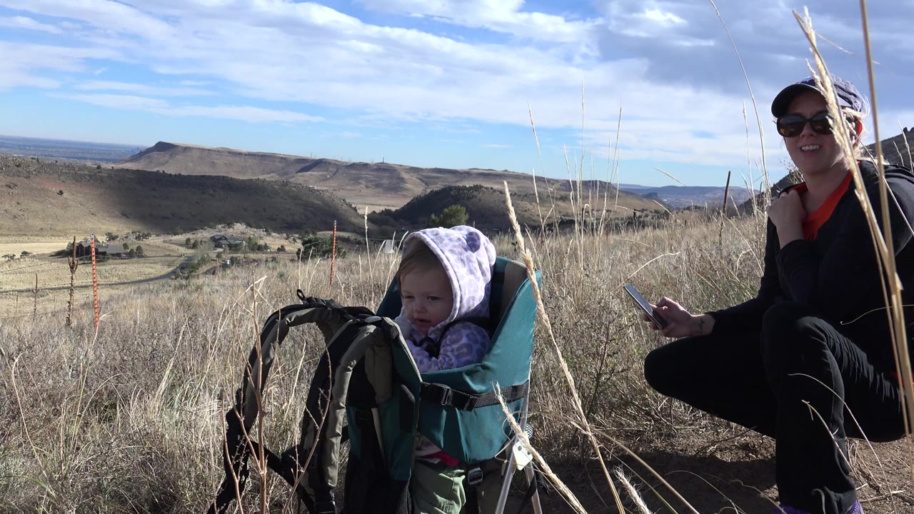 Baby in a hooded jacket looks forward while the mother sits smiling nearby.