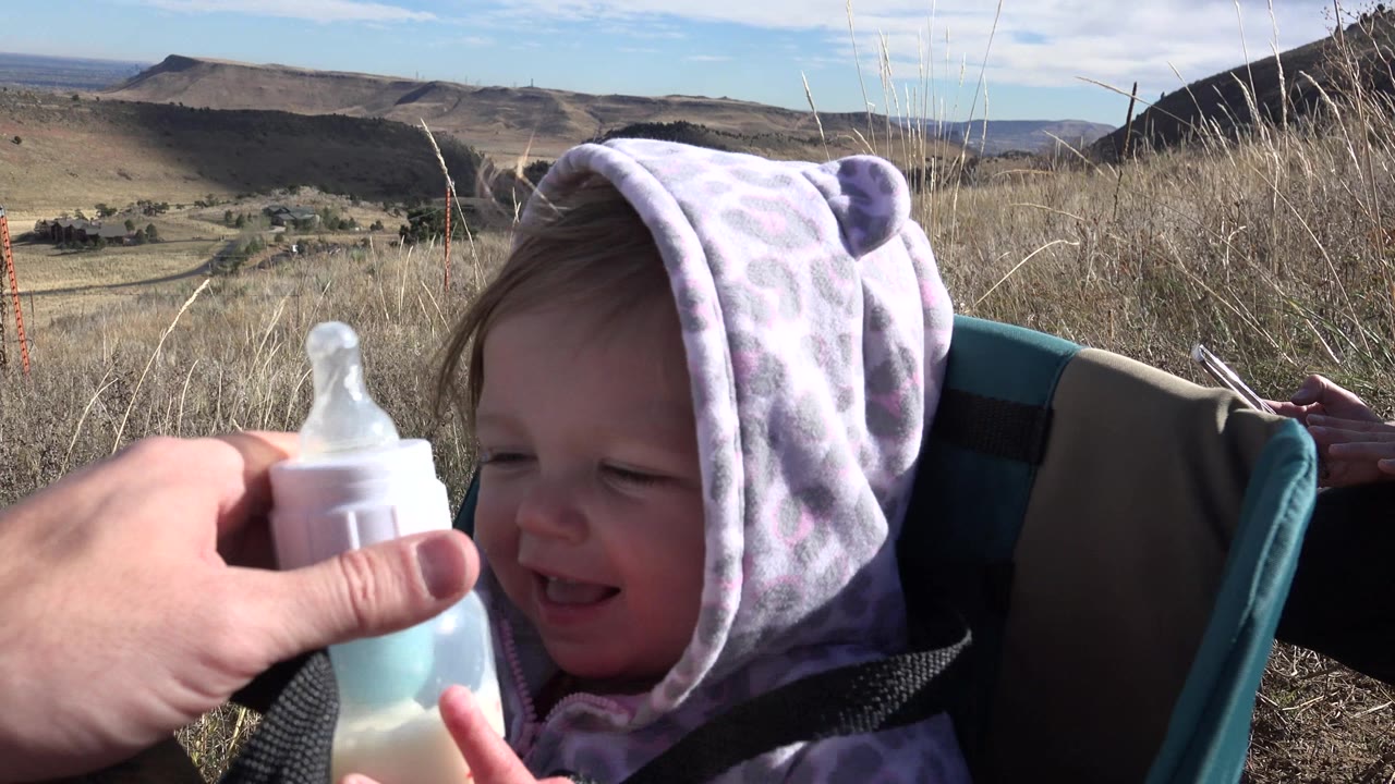 Baby in purple hood smiling at a bottle outdoors.