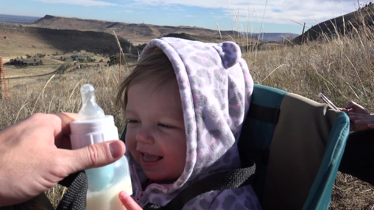 The baby sits in a teal and tan carrier and looks toward a bottle held close to their face in the Golden, Colorado landscape.