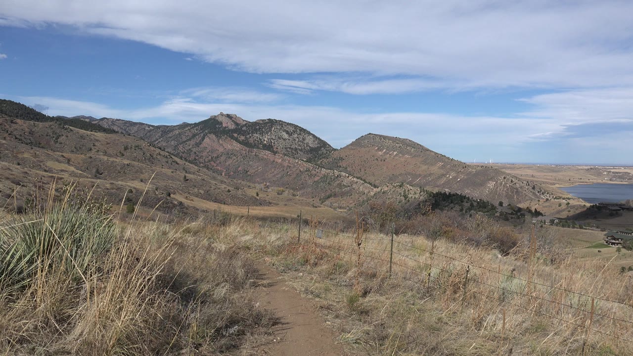 Opening view in Golden, Colorado of a dirt hillside trail, dry grasses, layered mountains, a small lake at right, and no visible people.