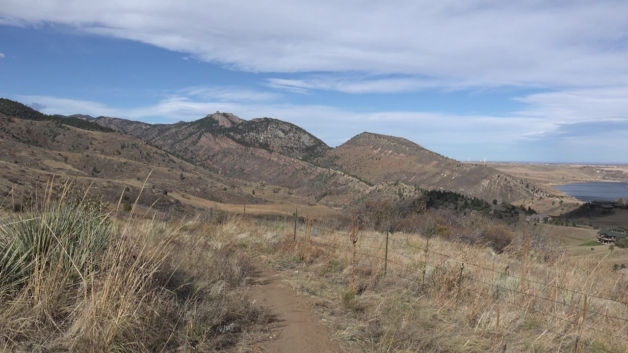 Golden, Colorado hillside view with taller dry grasses in the foreground, ridged mountains across the center, the lake near the right edge, and no visible people.