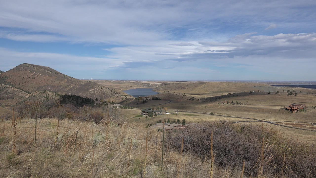 Golden, Colorado hillside view with dry foreground grass, brush, fence posts, a reservoir beyond the slope, scattered valley buildings, and no visible people.