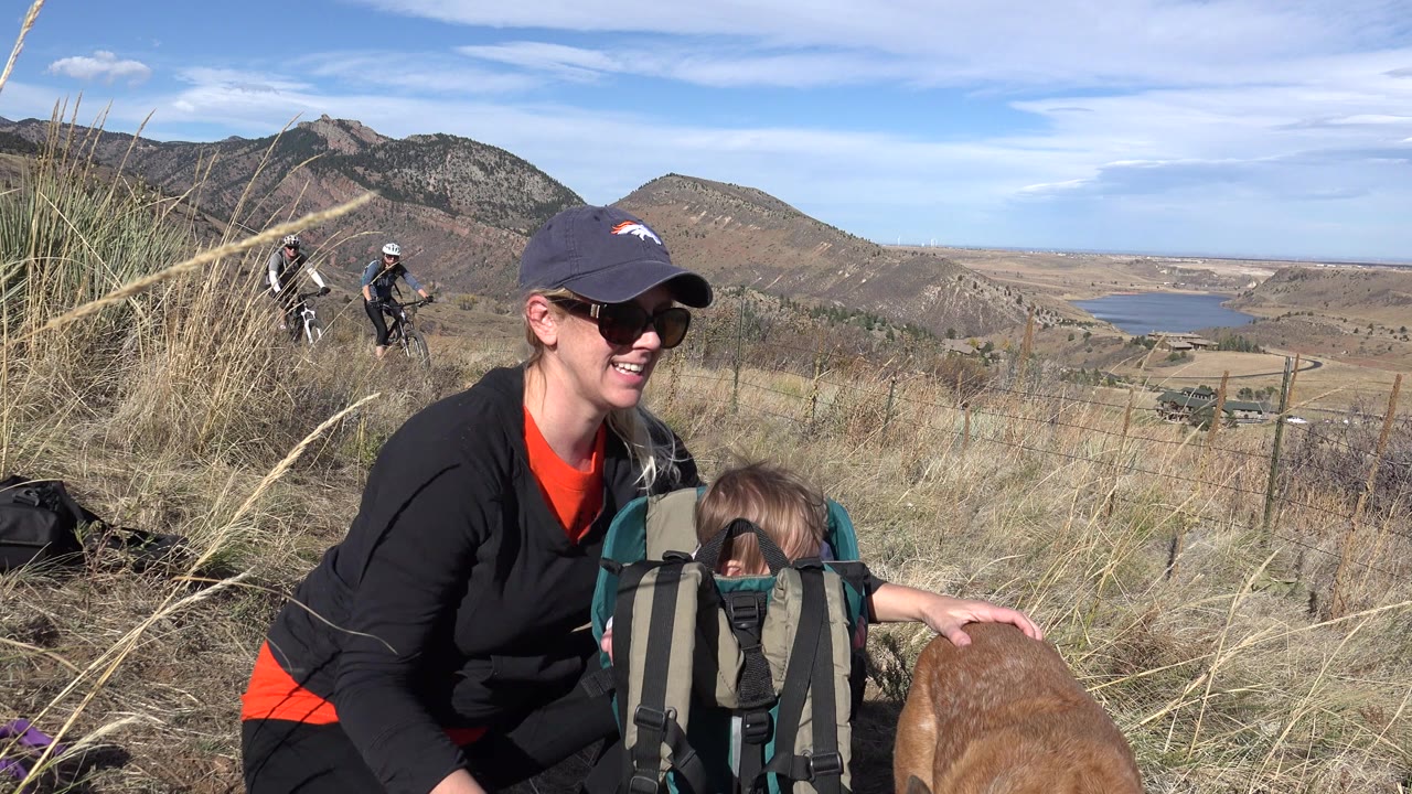 In Golden, Colorado, a woman in sunglasses kneels beside a child in a backpack carrier while a dog enters the lower frame and two cyclists appear on the hillside behind them.