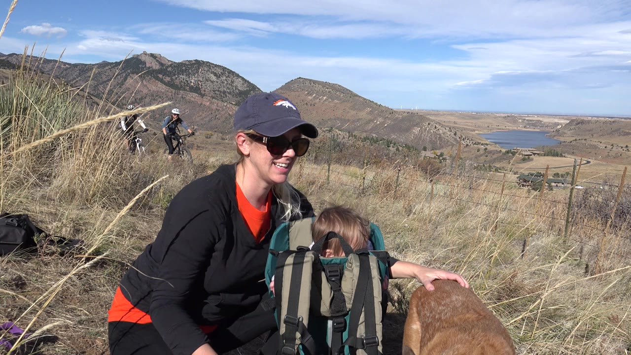 The woman smiles near the child carrier as the dog stays low at the edge of frame, with cyclists, dry grass, hills, and distant water visible in Golden, Colorado.