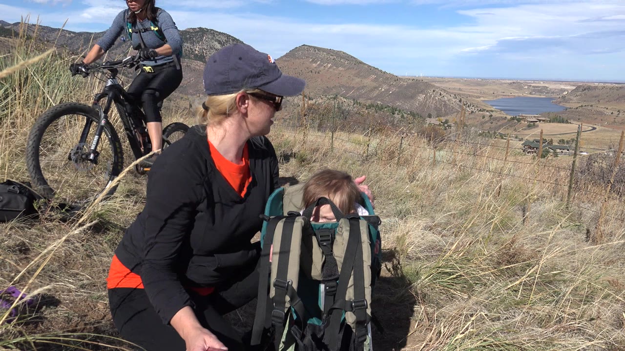 Mother seated with child in hiking backpack as a cyclist passes on a hillside trail.