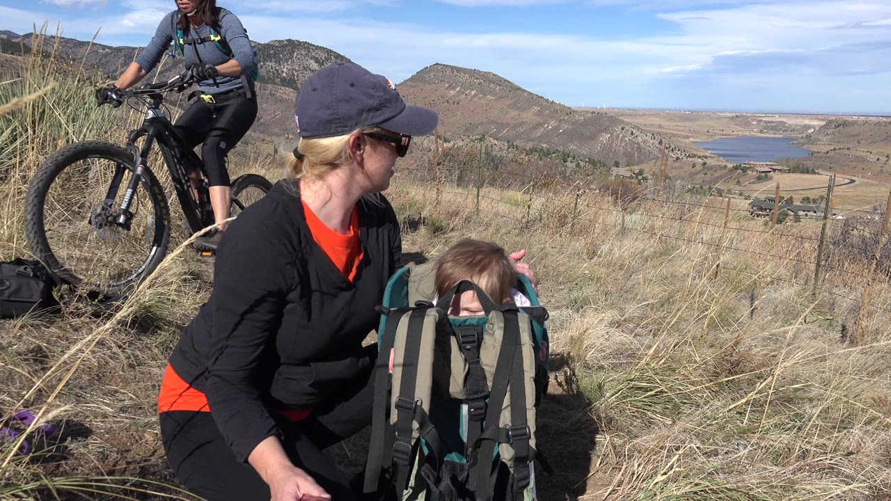 The mother remains turned toward the child in the hiking carrier in Golden, Colorado, with a close bicycle wheel at the left edge and the valley landscape stretching behind them.