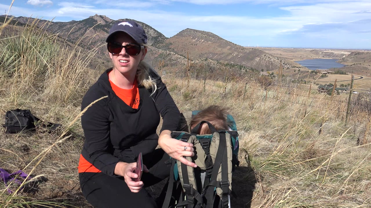Woman in sunglasses beside toddler backpack carrier on grassy hillside.
