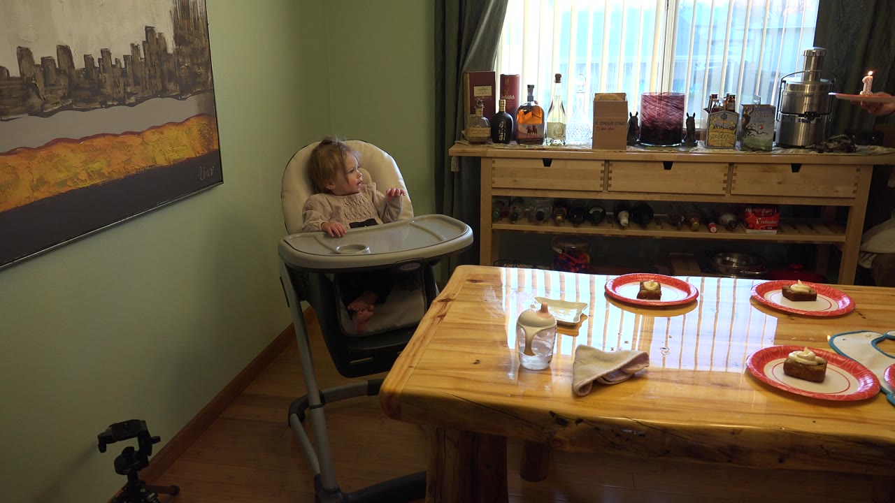 At 0:01 in Westminster, Colorado, the child looks toward a hand entering from the right near the table, with plates and a cabinet visible behind the foreground tabletop.