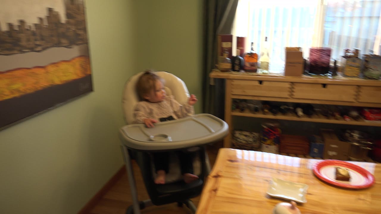 At 0:01 in Westminster, Colorado, the baby remains in the high chair beside the table while bright window light outlines bottles and household items on the shelf behind.