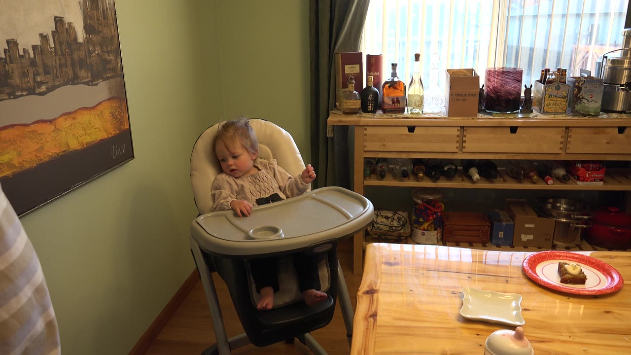 Young girl in a highchair beside a dining table with a cupcake on a red plate.