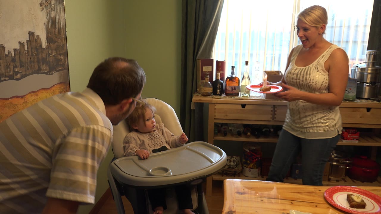 Adults and baby gathered near a high chair as a cake plate sits on the table.